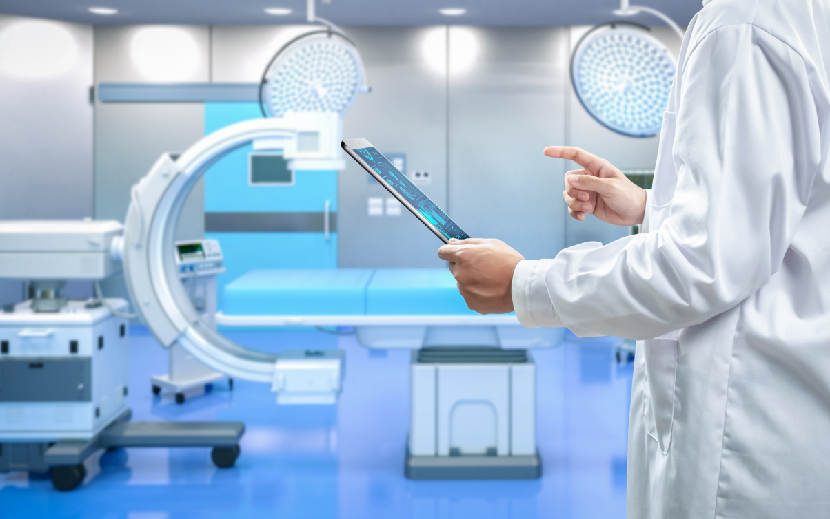 Photo of a doctor in a white lab coat reading results from a table in a hospital room with a C-Arm machine in the background